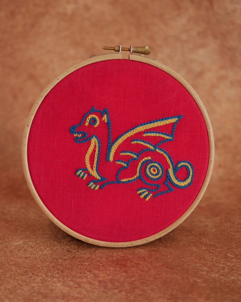 Broderie sur toile rouge d'un petit dragon mignon et naïf coloré dans un style médiéval. Au fil bleu et jaune.