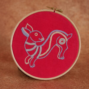 Broderie sur toile rouge : un lapin mignon et naïf en train de sauter. Il est brodé au fil bleu clair et beige.