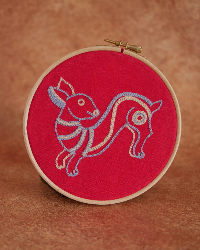 Broderie sur toile rouge : un lapin mignon et naïf en train de sauter. Il est brodé au fil bleu clair et beige.