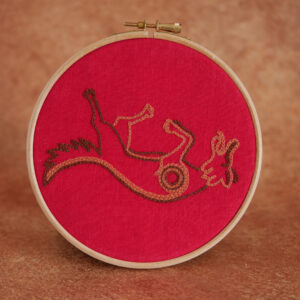 Broderie sur toile rouge : un renard couché sur le dos, la langue tirée. Il est brodé au fil marron et orange.