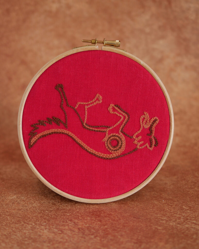 Broderie sur toile rouge : un renard couché sur le dos, la langue tirée. Il est brodé au fil marron et orange.