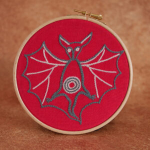 Broderie sur toile rouge : une chauve souris d'inspiration médiévale, les ailes écartées, de grandes oreilles et des yeux tout ronds. Elle est brodée au fil bleu gris et rose pâle.