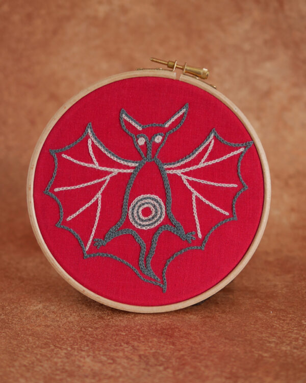 Broderie sur toile rouge : une chauve souris d'inspiration médiévale, les ailes écartées, de grandes oreilles et des yeux tout ronds. Elle est brodée au fil bleu gris et rose pâle.