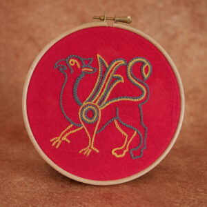 Broderie sur toile rouge : un griffon de profil avec un corps de lion, des ailes et une tête de rapace. Il est brodé au fil bleu gris et jaune.