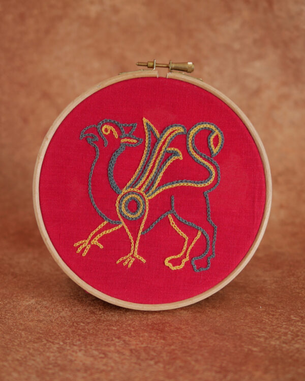 Broderie sur toile rouge : un griffon de profil avec un corps de lion, des ailes et une tête de rapace. Il est brodé au fil bleu gris et jaune.