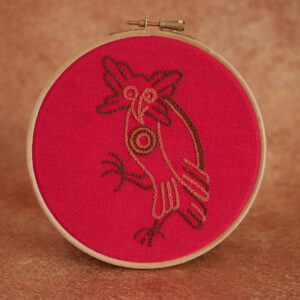 Broderie sur toile rouge d'une chouette/hibou médiévale brodée en marron et brun. Elle a l'air un peu bête, comme il se doit, le corps de profil, une patte en l'air et la tête tournée vers nous d'un air interrogateur.