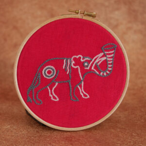Broderie sur tissu rouge : un éléphant naïf dans le style médiéval. Il a une trompe levée en trompette, une petite oreille en forme de feuille, une crinière ondulée et un air un peu perplexe. Il est brodé en bleu gris et rose pâle.