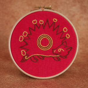 Broderie sur toile rouge : Un hérisson naïf inspiré des enluminures médiévales. Il est marron, de profil avec des fruits jaunes plantés sur ses épines. Il a une tête mignonne, on dirait qu'il sourit et une petite queue en tire-bouchon.