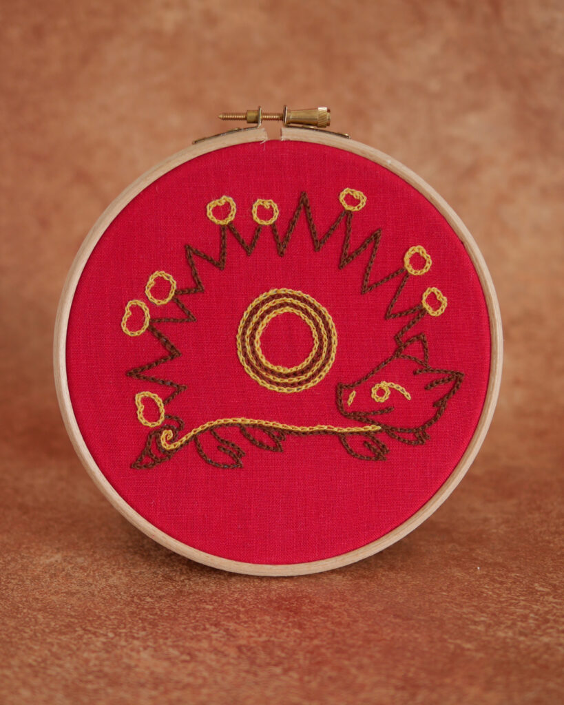 Broderie sur toile rouge : Un hérisson naïf inspiré des enluminures médiévales. Il est marron, de profil avec des fruits jaunes plantés sur ses épines. Il a une tête mignonne, on dirait qu'il sourit et une petite queue en tire-bouchon.