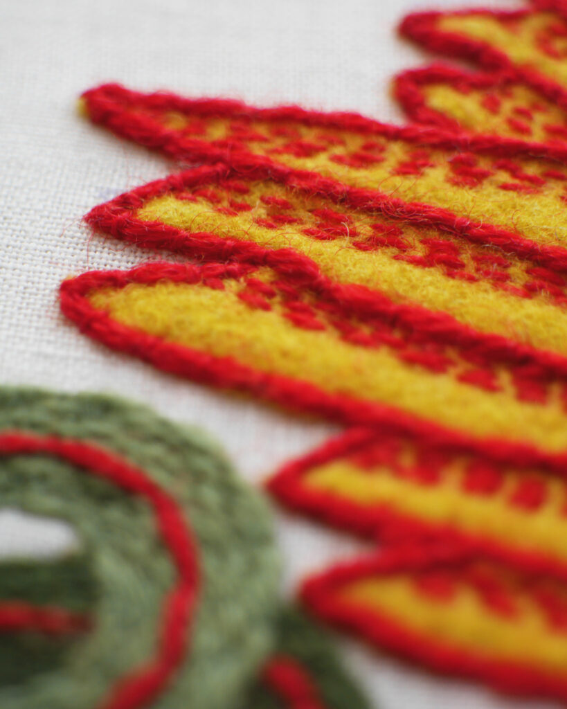 Gros plan sur l'aile en tissu jaune cousue sur toile blanche avec un point de broderie rouge sur le contour et des petits points rouges pour rehausser au centre.