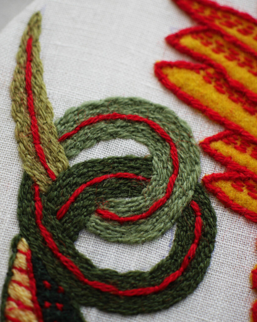 Gros plan sur la broderie de la queue, qui s'enroule sur elle-même en forme de bretzel. La queue est brodée en nuances de vert de plus en plus clair vers la pointe. Avec une ligne rouge au centre.
