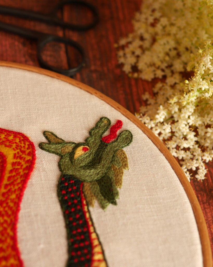 Gros plan sur la tête d'un dragon brodé. Il a une barbe, un œil rouge et la lange tirée. Il regarde vers un bouquet de fleurs de sureau posé sur la table.