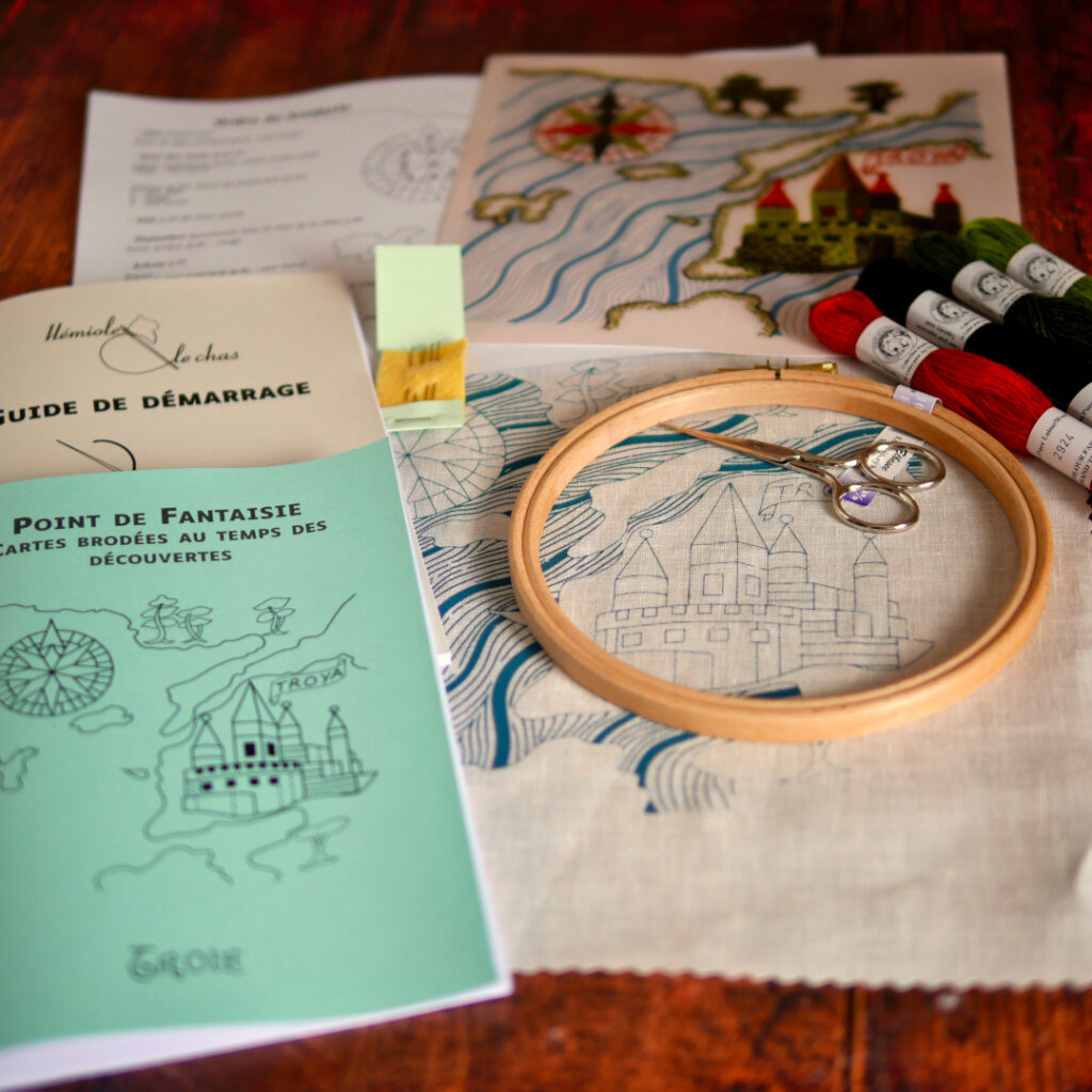 Contenu du kit posé sur une table : la toile imprimée, 4 échevettes de laine colorées, 4 aiguilles, un tambour, une paire de ciseaux, 2 livrets imprimés, une impression couleur du modèle.