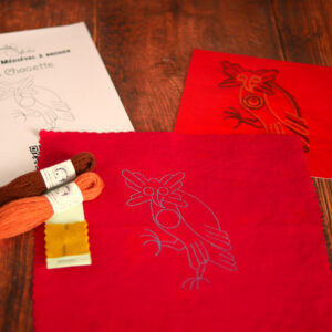 Du matériel de broderie posé sur une table : une toile rouge imprimée avec une chouette médiévale, un livret et une photo couleur de la broderie, 2 échevettes de laine colorée et une aiguille de broderie.