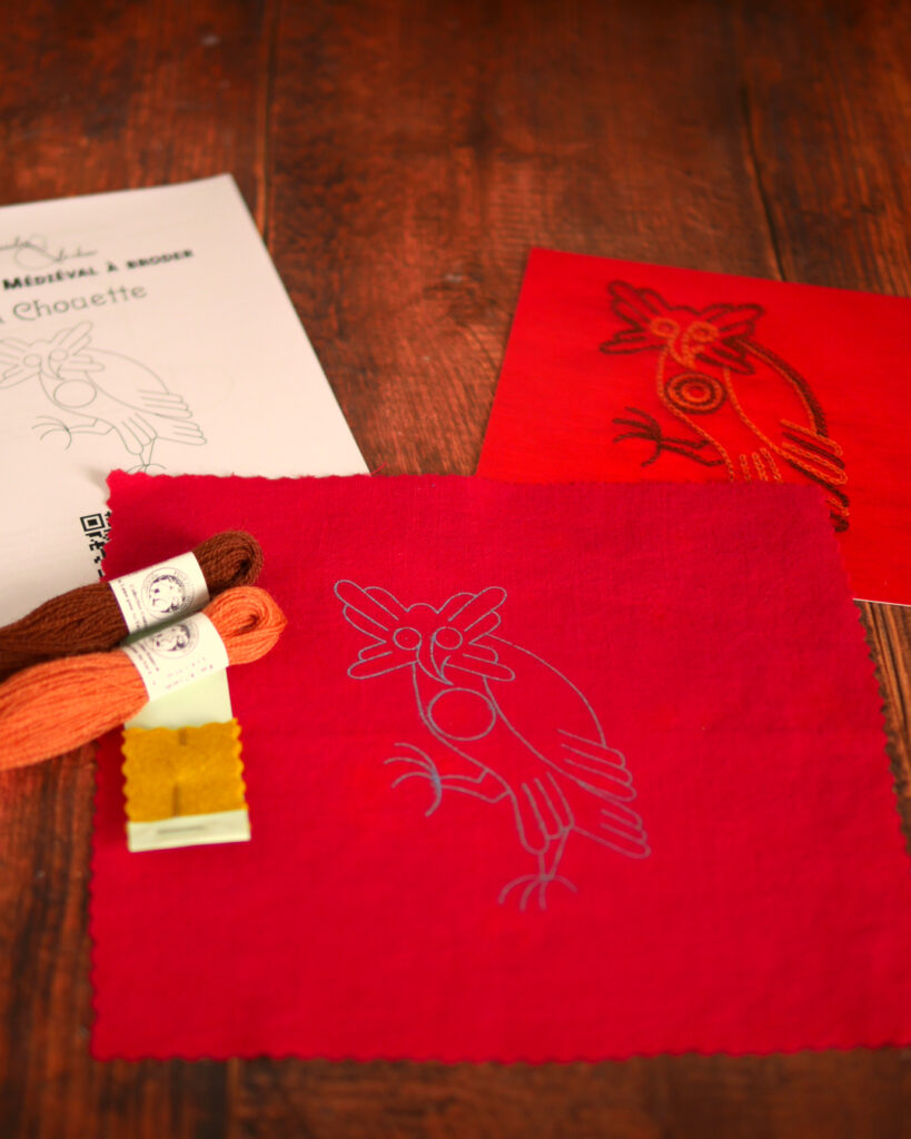 Du matériel de broderie posé sur une table : une toile rouge imprimée avec une chouette médiévale, un livret et une photo couleur de la broderie, 2 échevettes de laine colorée et une aiguille de broderie.
