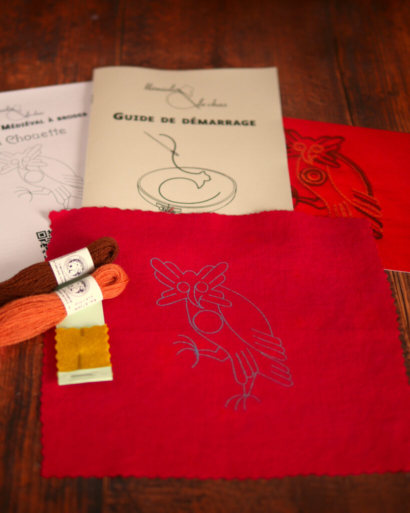 Du matériel de broderie posé sur une table : une toile rouge imprimée avec une chouette médiévale, un livret et une photo couleur de la broderie, 2 échevettes de laine colorée et une aiguille de broderie, un livret imprimé où il est écrit "guide de démarrage".