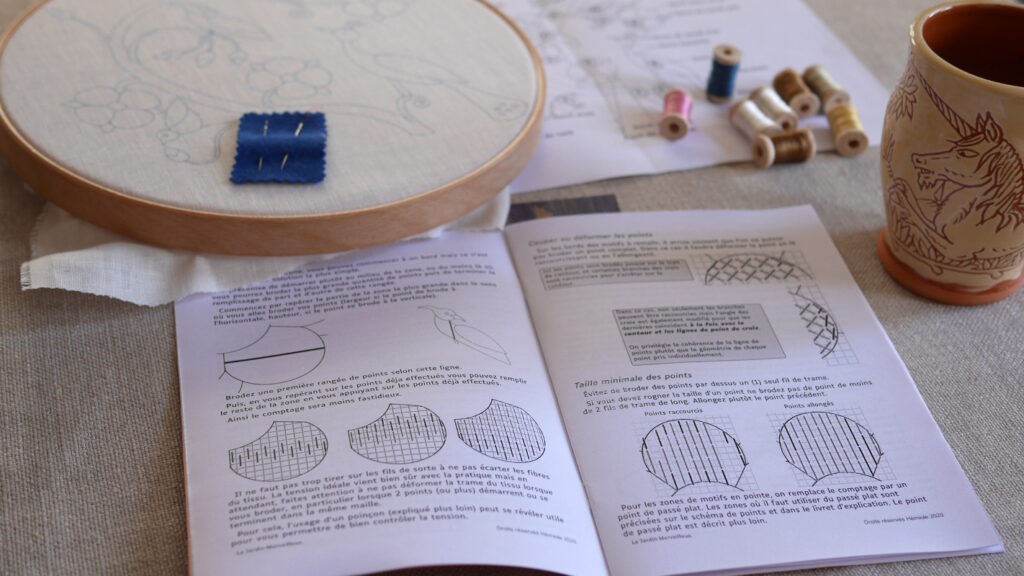 Un livret imprimé ouvert sur une page où on voit du texte est des schémas. Au dessus un tambour de broderie avec une toile blanche imprimée, des bobines de fils pastels, des aiguilles.