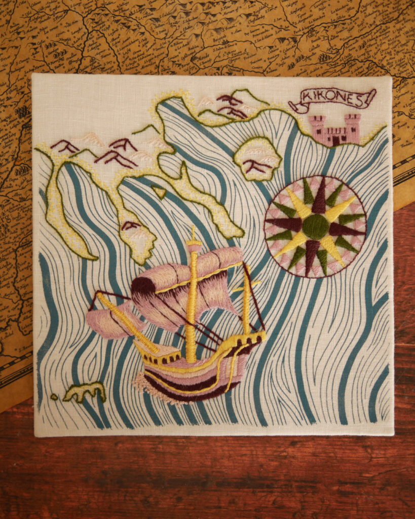 Broderie d'une carte marine les côtes avec des montagnes et du sable, une rose des vents brodée en jaune vert rose et pourpre, un bateau style caravelle brodé en rose jaune et pourpre avec un peu d'écume devant. La mer est imprimée en vagues bleues.