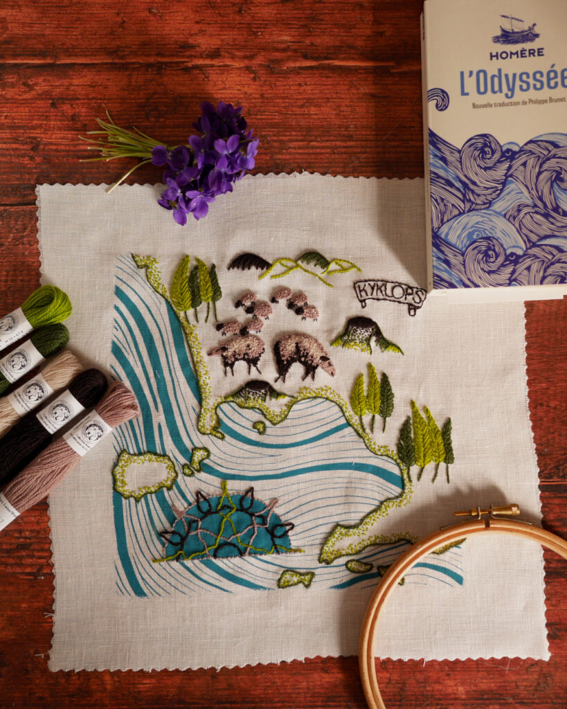Une broderie posée sur une table. Il s'agit d'une carte où la mer est imprimée avec des vagues bleues. La côte est bordée de plusieurs îles brodées en vert. Une demi rose des vents en motifs d'entrelacs sur fond bleu. Dans les terres, il y a des bosquets de peupliers, des collines, des volcans et des moutons bruns. Un cartouche en forme de parchemin où il est écrit "KYKLOPS".