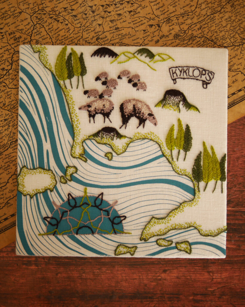 Une broderie posée sur une table. Il s'agit d'une carte où la mer est imprimée avec des vagues bleues. La côte est bordée de plusieurs îles brodées en vert. Une demi rose des vents en motifs d'entrelacs sur fond bleu. Dans les terres, il y a des bosquets de peupliers, des collines, des volcans et des moutons bruns. Un cartouche en forme de parchemin où il est écrit "KYKLOPS".