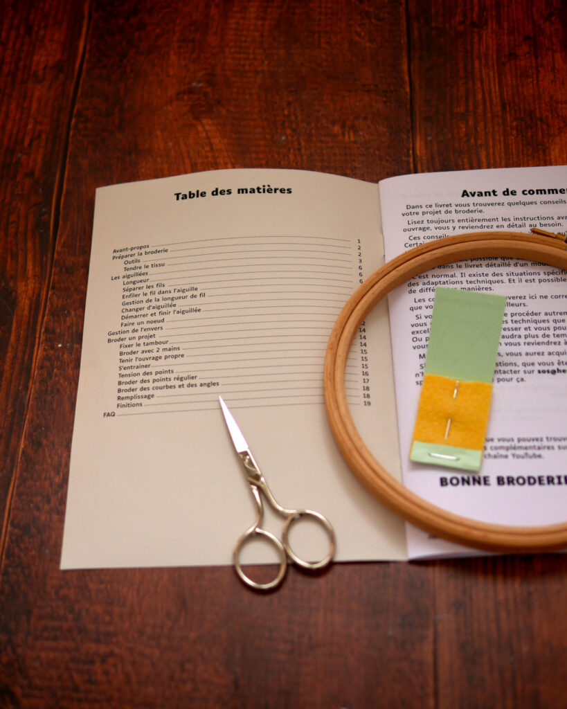 Un livret ouvert à la page de la table des matières. Par dessus, un petit tambour de broderie en bois, une aiguille de broderie et une petite paire de ciseaux de broderie.