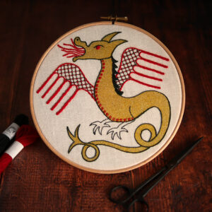 Un dragon brodé dans un tambour de broderie. Il a le corps et la tête imprimés en aplat d'encre dorée et les contours, les ailes et quelques détails sont brodés aux fils vert foncé et rouge Les dragons d'arrière plan sont brodés avec des couleurs vives : différentes nuances de vert, jaune et rouge.