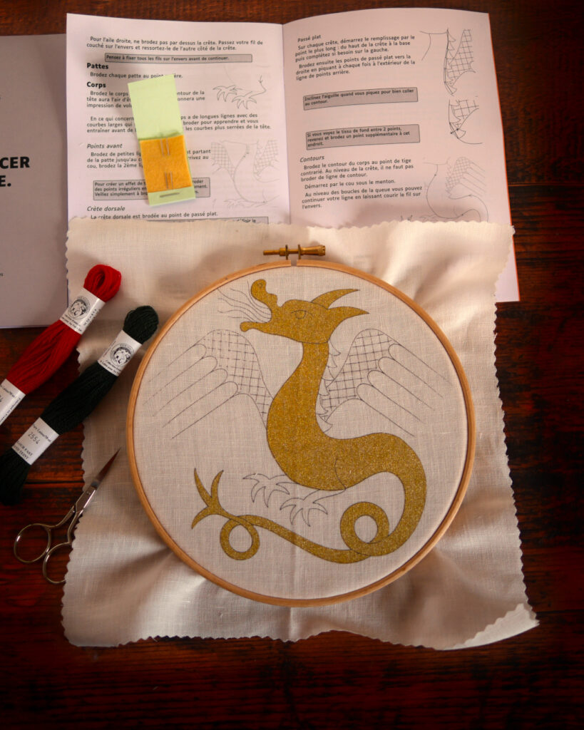 Tambour de broderie et matériel posés sur une table. Dans le tambour, une toile imprimée avec un dragon rempli à l'encre dorée.