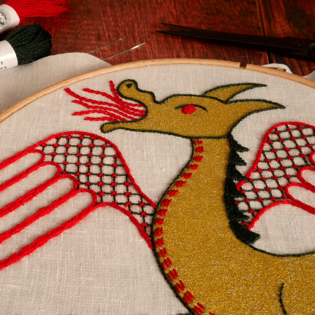 Détail de la broderie du dragon doré : on voit le haut du corps et la tête Les différentes parties sont brodées au fil vert foncé et rouge avec des points différents pour chaque zone : un treillis sur les ailes, une crête en aplat vert sur le dos, des chaînettes pour les flammes qui sortent de sa gueule et quelques petits points avant pour souligner la ligne du ventre.
