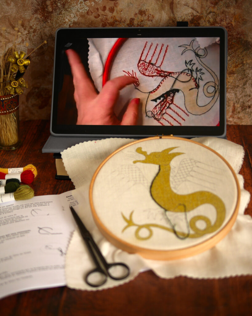 Sur une table, une broderie en cours est posée dans son tambour. C'est un dragon médiéval dont le corps est imprimé à l'encre dorée. La broderie est commencée par un contour vert foncé. Derrière, il y a une tablette posée avec une vidéo en cours de lecture. On reconnait le dragon en train d'être brodé.