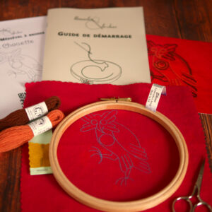 Du matériel de broderie posé sur une table : une toile rouge imprimée avec une chouette médiévale, un livret et une photo couleur de la broderie, 2 échevettes de laine colorée et une aiguille de broderie, un livret imprimé où il est écrit "guide de démarrage", un tambour de broderie et des petits ciseaux.