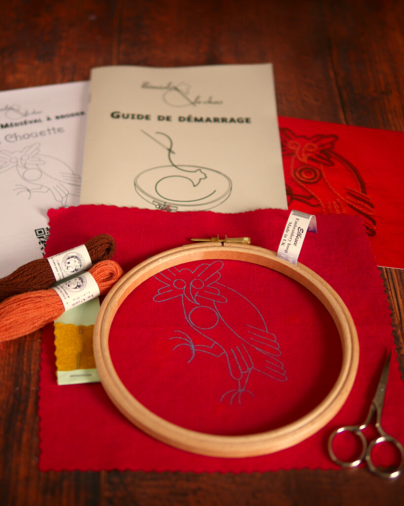 Du matériel de broderie posé sur une table : une toile rouge imprimée avec une chouette médiévale, un livret et une photo couleur de la broderie, 2 échevettes de laine colorée et une aiguille de broderie, un livret imprimé où il est écrit "guide de démarrage", un tambour de broderie et des petits ciseaux.