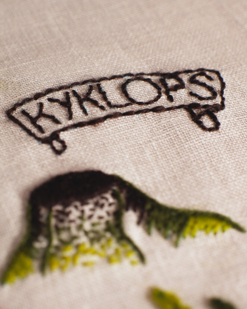 Gros plan sur une broderie sur toile blanche. Un phylactère en forme de parchemin dans lequel est écrit "KYKLOPS" et en avant plan, un volcan aux pentes vertes.