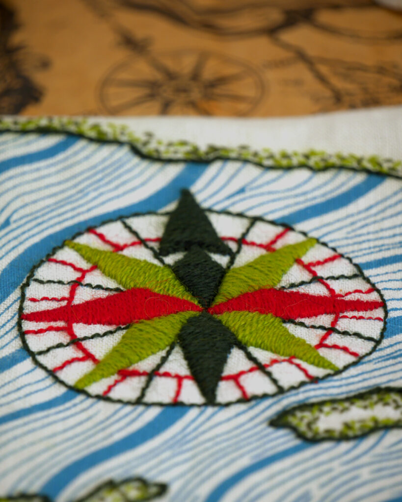 Détail de la broderie : la rose des vents. Les grandes flèches des directions principales sont brodées en vert foncé, rouge et vert clair. Elles sont inclues dans des cercles rouge et vert foncés avec des lignes fines indiquant les directions intermédiaires. En arrière plan on devine la rose des vents de la carte ancienne sur laquelle la broderie est posée.