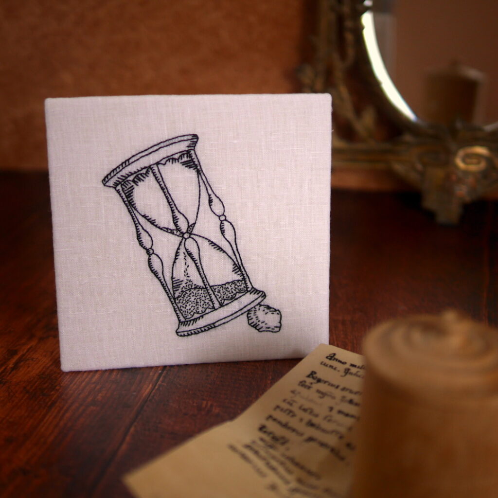 Broderie au fil noir sur toile blanche dans le style des gravures de la renaissance Elle représente un sablier, posé penché sur un cailloux.