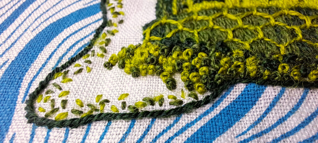 Détail d'une broderie de carte marine en gros plan : On distingue une côte brodée au point de sable avec la mer imprimée à l'encre bleue. Dans les terres, on voit le bas d'une muraille et des rochers brodés au point de nœud en différentes nuances de vert.