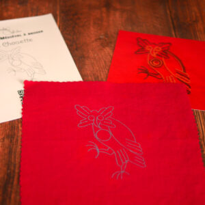 Du matériel de broderie posé sur une table : une toile rouge imprimée avec une chouette médiévale, un livret et une photo couleur de la broderie.