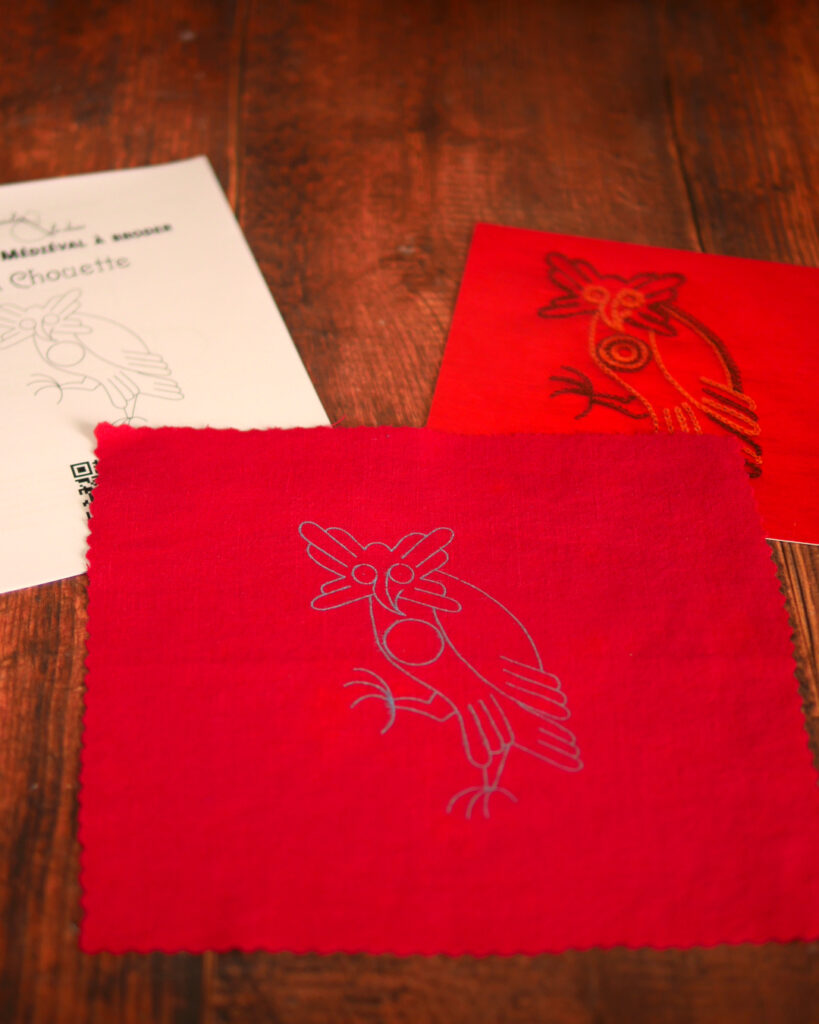 Du matériel de broderie posé sur une table : une toile rouge imprimée avec une chouette médiévale, un livret et une photo couleur de la broderie.