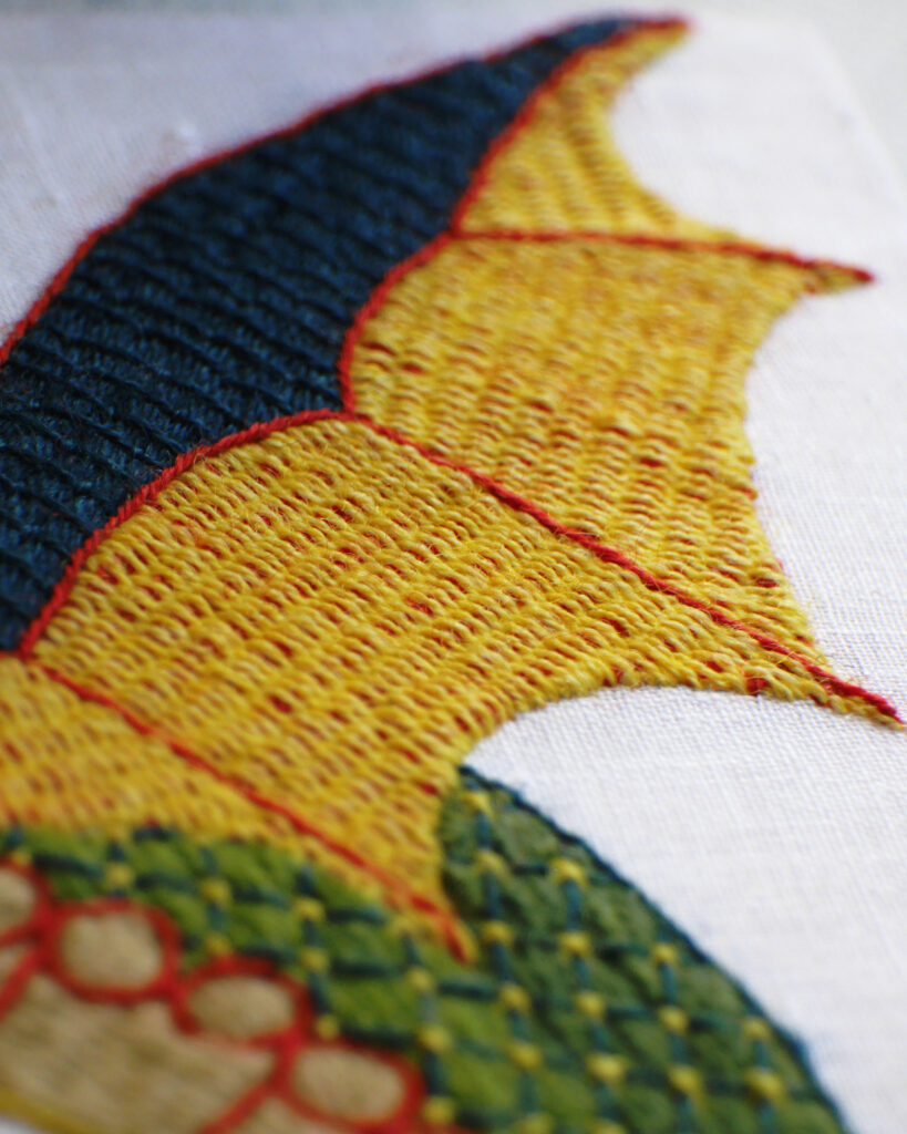 Détail de la broderie d'une aile du dragon. Elle est en 2 parties : le haut est brodé en vert foncé. Le bas qui est découpé comme des ailes de chauve-souris est brodé en jaune avec du rouge qui apparait en dessous.