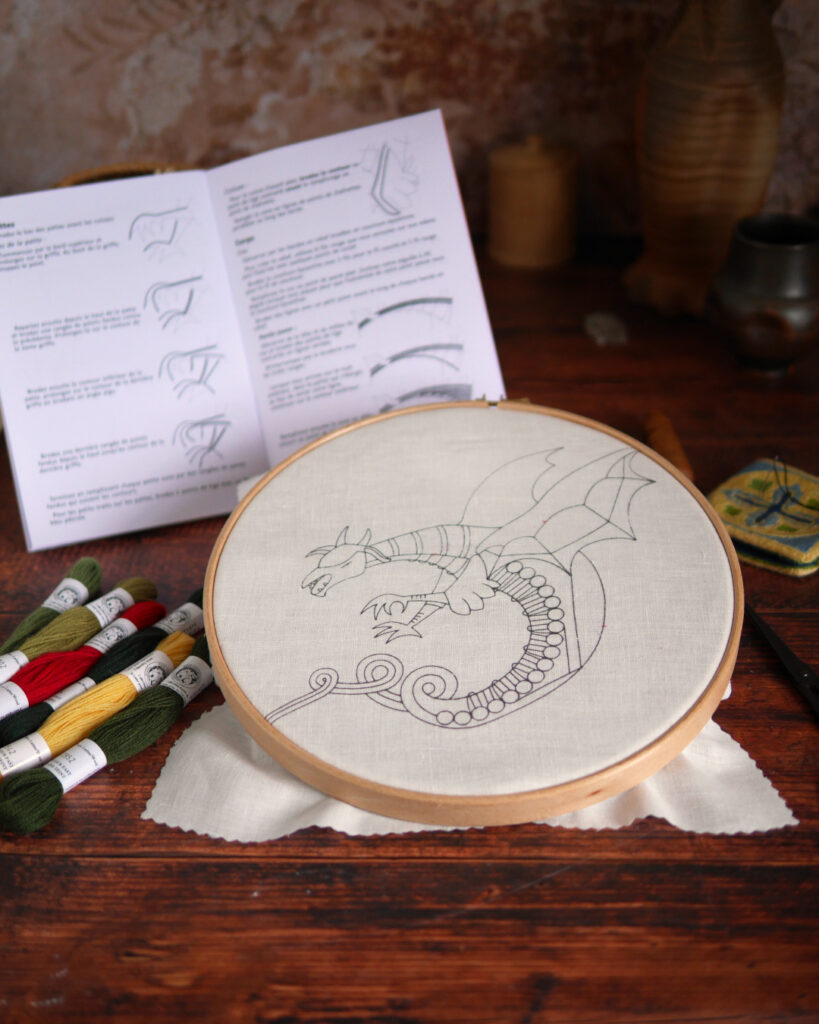 Une table où sont posés des affaires de broderie Au centre, un tambour de broderie avec une toile où un dragon est imprimé A côté, des échevettes de fils colorés et un livret avec du texte et des dessins.