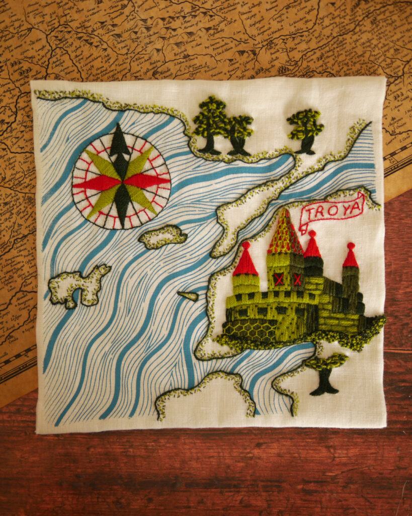Une carte marine brodée de la côte Turque au niveau la ville de Troie. La toile de lin blanc est posée sur une table sur une carte ancienne avec quelques coquillages, un verre à pied et du bazar. La mer, représentée par des vagues bleues est imprimée. La côte, ainsi qu'une rose des vents et une cité fortifiée sont brodées en différentes nuances de vert et du rouge. Le long de la côte, des petits points sont brodés et quelques arbres au feuillage chaotique.