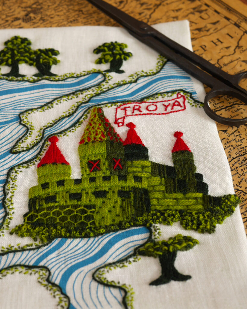 Détail de la broderie centré sur la cité brodée en vert avec des toits rouges sur plusieurs tours. Au dessus un cartel en forme de parchemin où il est écrit "Troya".