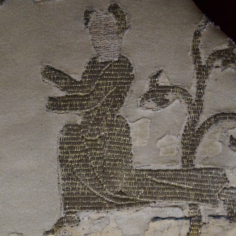 Gros plan sur une broderie médiévale abîmée. Sur toile blanche, un personnage agenouillé, derrière lui, un arbre.
La broderie est réalisée au fil d'or couché verticalement avec une grande régularité. Les points de fixation forment des lignes horizontales.
Les points de couchure sont tirés sur l'envers de la broderie et donnent l'impression que le fil d'or est brodé au travers du tissu.