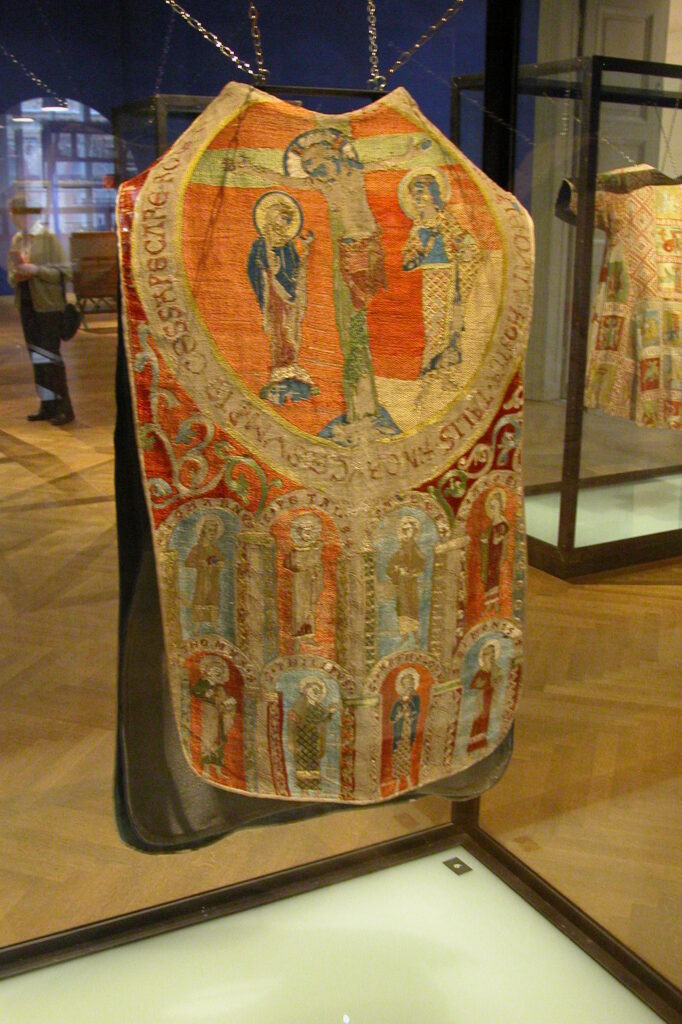 Chasuble entièrement brodée en soie colorée avec une dominante rouge (orangé).
Sur la partie haute, dans un grand cercle il y a un christ en croix entouré de 2 femmes. autour un texte.
Sur la partie basse dans un décors d'arches, sur 2 rangées de 4 : différentes figures de saints sur fond rouge et bleu alternés.