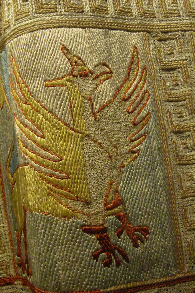Détail d'un animal brodé de la tunique. Un griffon brodé en jaune sur fond bleu clair avec le contour, les pattes et l’œil rouge.
La broderie est réalisée en petits points verticaux parfaitement réguliers qui forment une légère texture diagonale.
