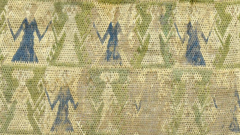 Détail d'une broderie sur fond vert et jaune, des personnages bleu et blanc en ligne dans une position de danse, comme s'ils se tenaient les mains.
C'est brodé en petits points et très schématiques.