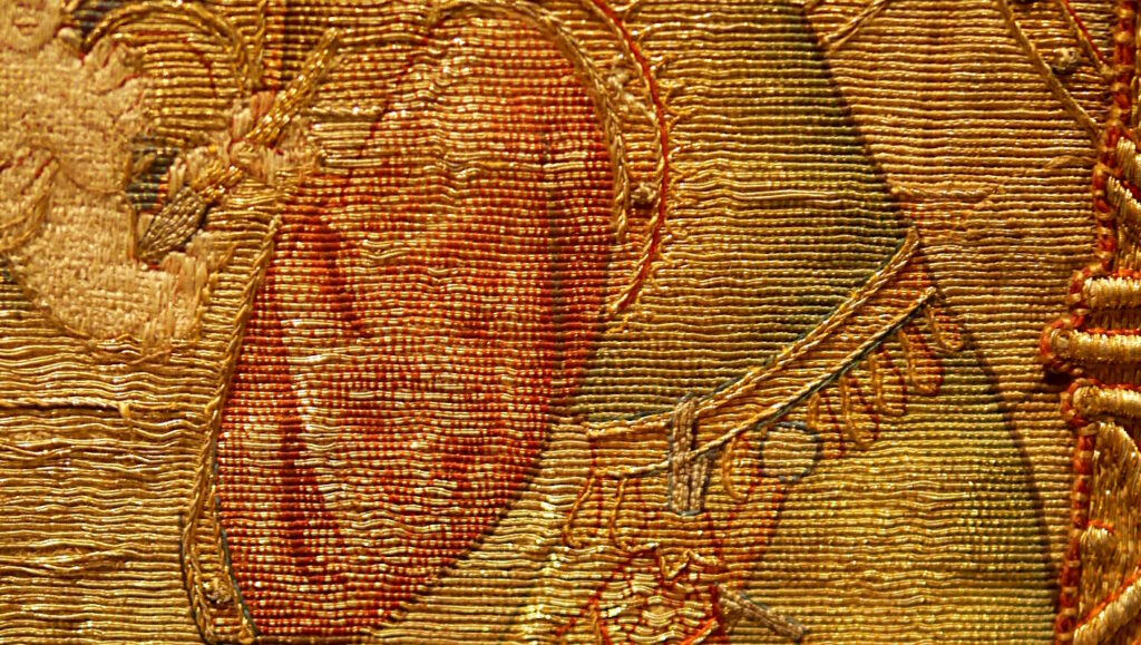 Gros plan sur la broderie en or du buste d'un homme.
La broderie est entièrement recouverte de fil d'or couché horizontalement.
Les points de fixation sont brodés avec des fils colorés.
Les fils colorés de couchure, de différentes couleurs, sont brodés de manière plus ou moins espacés pour laisser apparaître le fil d'or.