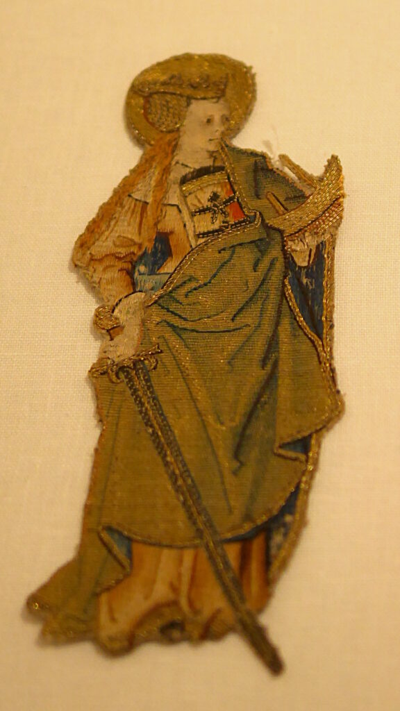 Broderie médiévale. Une sainte brodée en or et soie.
Elle tient une épée dans une main et un bateau dans l'autre.