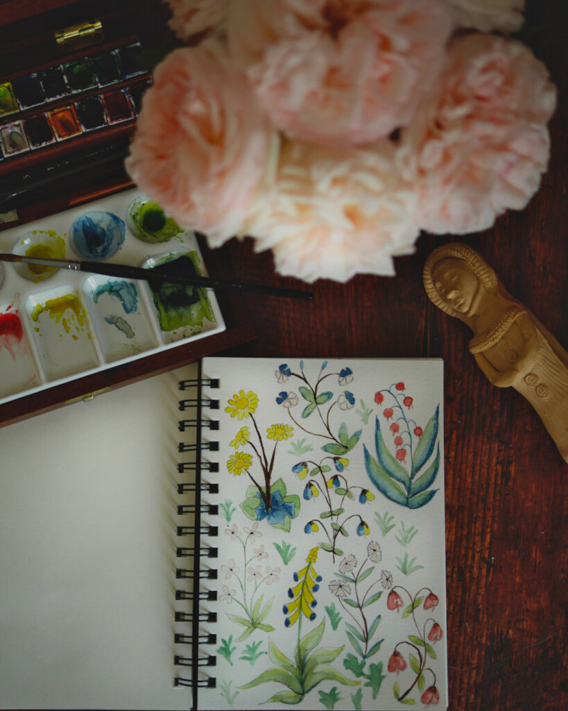 Une table sur laquelle est posé un carnet et de l'aquarelle.
sur le carnet sont peintes différentes fleurs dans un style plus ou moins médiéval.
Un bouquet de roses roses pâles est posé devant.