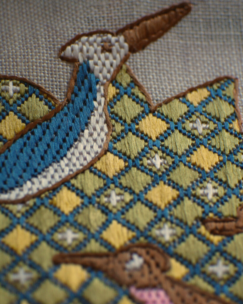 Gros plan sur une broderie dans le style médiéval sur toile blanche brodée avec des fils colorés qui forment des petits points géométriques. Le feuillage d'un arbre, brodé avec un damier vert avec des bordures bleues. Dans le damier, certains carreaux sont jaunes et d'autres ont une petite croix blanche au centre. Il y a 2 oiseaux brodés par dessus : un bleu et blanc en haut et un marron et rose en bas.