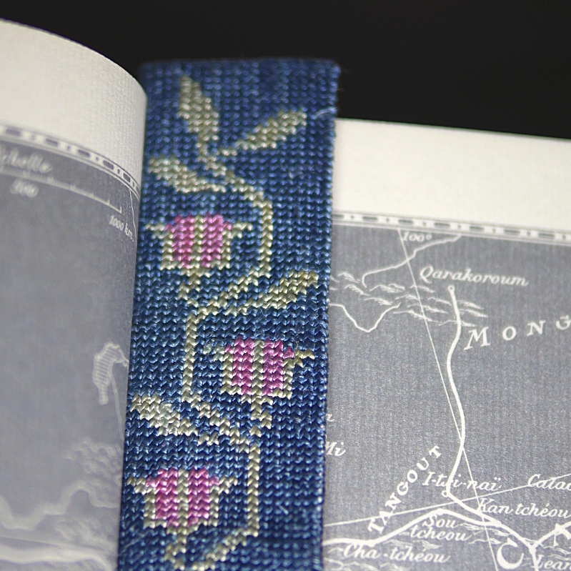 Un marque page brodé dans un livre ouvert. La broderie est une frise fleurie sur fond bleu, avec des roses à peine écloses.
